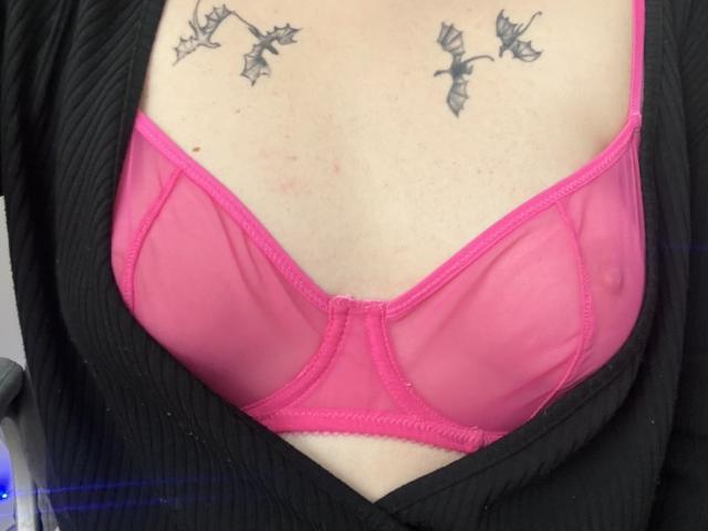 Rubbi's pink bra is sheer, showing her tits, with black ribbed cardigan open. Rubbi has tattoos of bats above her tits. Her skin is light.