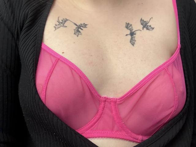 Rubbi's pink bra shows her tits, with black tattoos of flowers on her chest. She's wearing a black ribbed cardigan. Her skin is light.