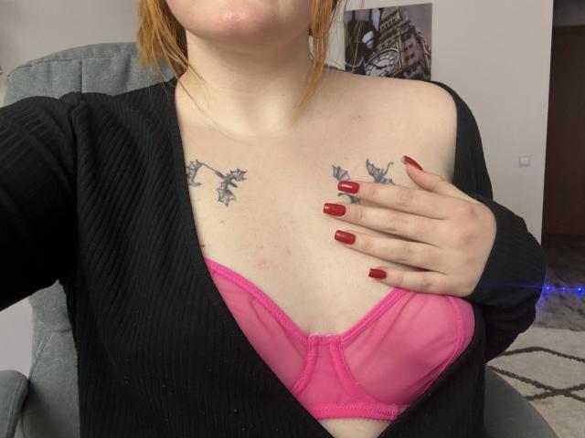 Rubbi's tits are in a pink bra, with black tattoos on her chest, red nails, and a black cardigan, showing her right hand over her left breast.