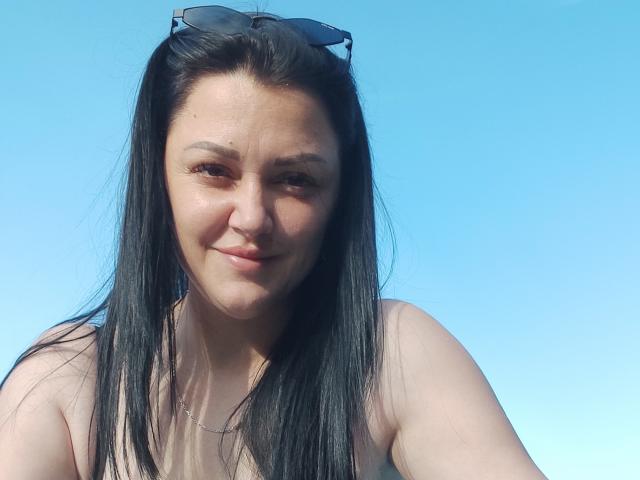 XCrazyBabeX with long black hair, sunglasses on head, slight smile, light brown skin, wearing a sleeveless top, clear blue sky background, looking at camera, slight shadow on face.