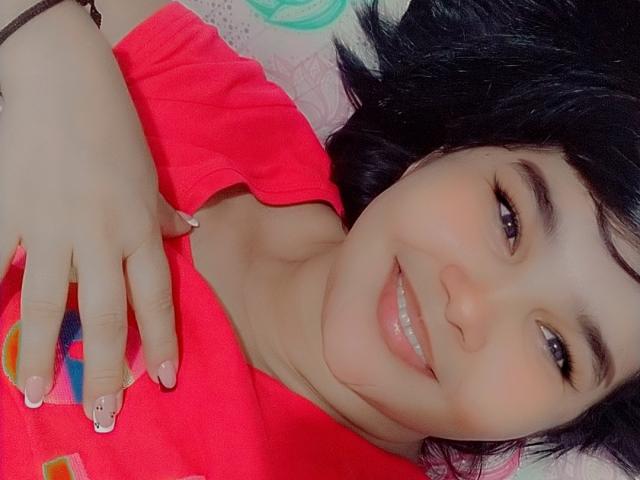 PrittyAlicia with black hair, smiling, wearing a red shirt, lying down, showing off her cute, small tits, and painted nails with a tiny face design.