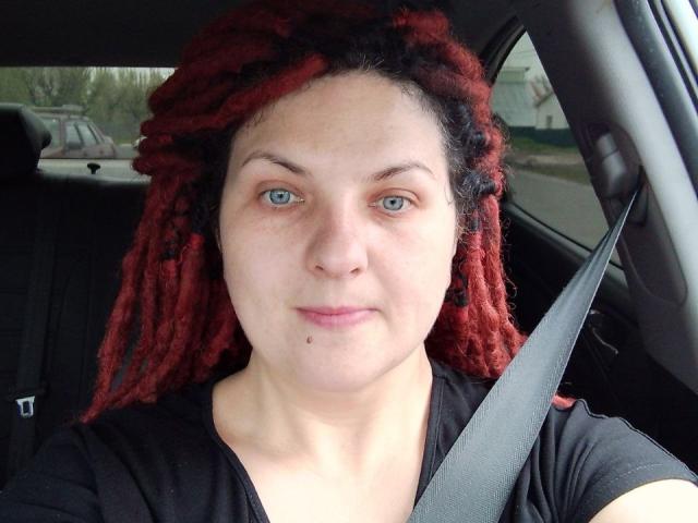 TaisiawSolo has red and black dreadlocks, blue eyes, a small mole under her nose, and wears a black shirt with a seatbelt across her chest in a car.