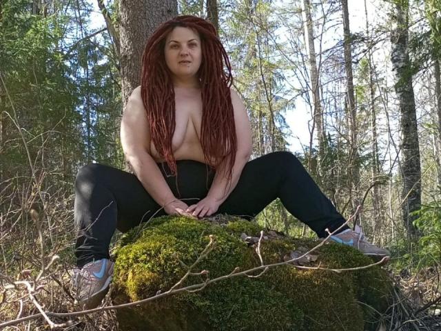 TaisiawSolo, topless with long red dreadlocks, sitting on a mossy rock in a forest, wearing black pants and sneakers, with her tits and ass exposed, looking at the camera.