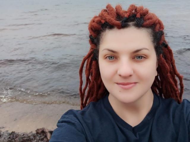 TaisiawSolo with red and black dreadlocks, light skin, blue eyes, wearing a dark blue shirt, standing by the water with a small wave touching the shore.