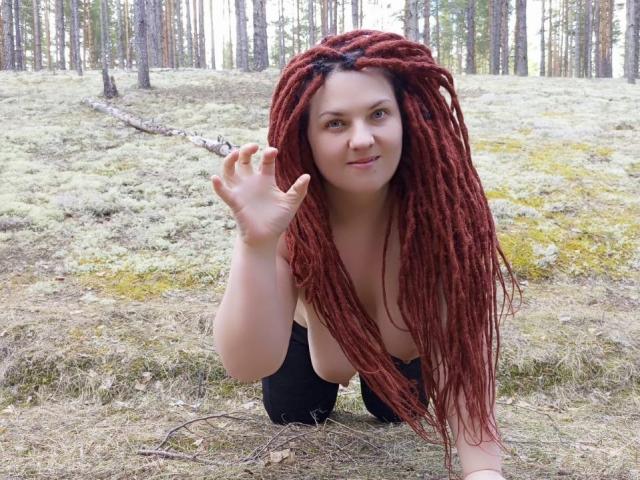 TaisiawSolo with long red dreadlocks, kneeling in a forest, waving with her right hand, wearing black pants, showing her tits, smiling slightly, fair skin, green eyes.
