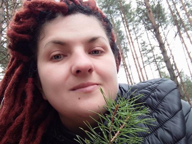 TaisiawSolo has red dreadlocks, a black puffy jacket, and a small mole under her nose, holding a pine branch with green needles near her face.