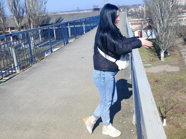 XCrazyBabeX with long black hair, black puffer jacket, blue jeans, white sneakers, and a white crossbody bag, leaning on a blue railing, looking to the right.