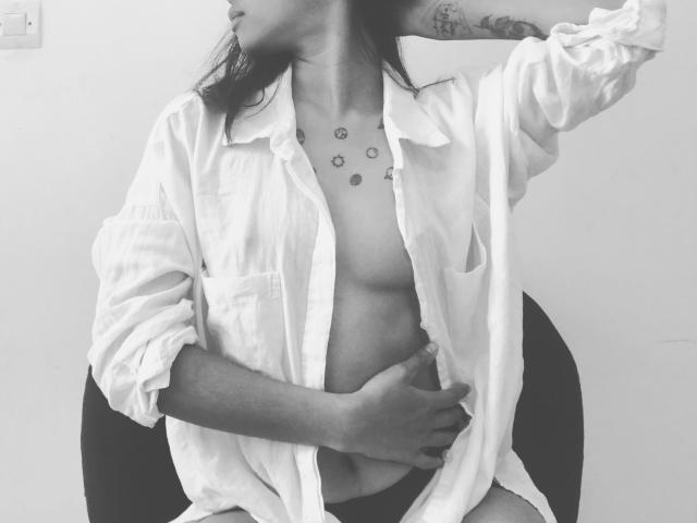 Foto em preto e branco de Omaliia com a camisa aberta, uma mão no abdômen, a outra no cabelo, seios pequenos visíveis, tatuagem no pescoço, sentada em uma cadeira, olhando para o lado..