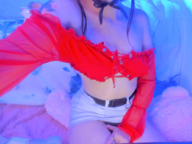 FairyBeauti portant un haut rouge à une épaule, avec des boutons qui laissent entrevoir de petits seins, un short blanc, une ceinture noire et un ruban noir, assise dans une pièce éclairée en rose et bleu..