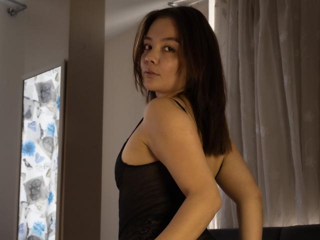 AmyCalloway with shoulder-length brown hair, wearing a black sheer top, looking over her shoulder, her ass slightly visible, standing in a dimly lit room.