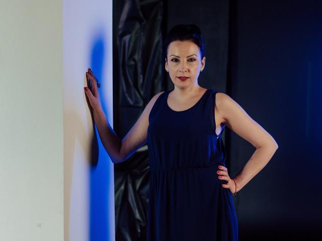 Elizabeth staat met één hand op een muur, ze draagt een donkerblauwe jurk, haar hand met rode nagels rust op haar heup, ze heeft kort zwart haar, een lichte huid en een zelfverzekerde glimlach..