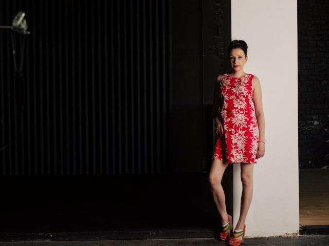 ElizabethSo stands against a white column, wearing a red floral dress, showcasing her tits and ass, with colorful heels, dark hair in a bun, and a serious expression.