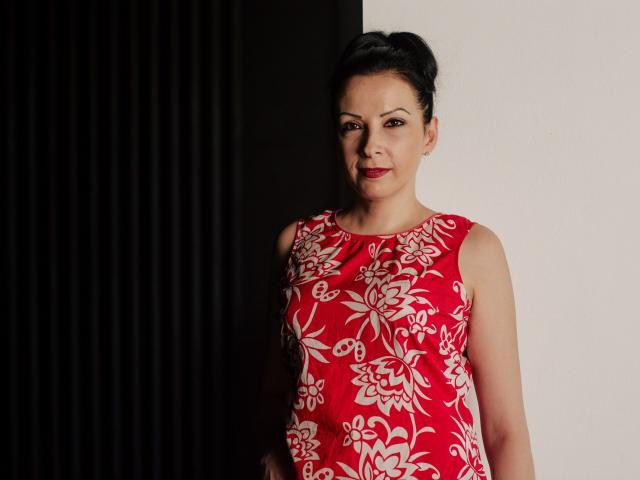 ElizabethSo has dark hair in an updo, wearing a red floral dress, standing with a confident look, her tits modest, red lipstick, against a black and white wall.
