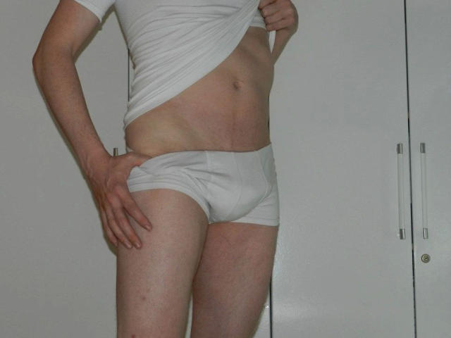 A white-tshirt-wearing, white-briefs-wearing, slim, light-skinned, hot dude lifts his shirt to show his flat, toned abs and slight hip bones, with one hand on his butt.