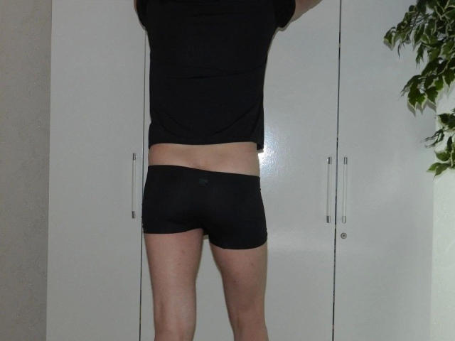 black t-shirt, black boyshorts, bare midriff, white wardrobe, plant in corner, standing with arms up, butt, legs, flat stomach, light skin, subtle muscle definition, no face visible