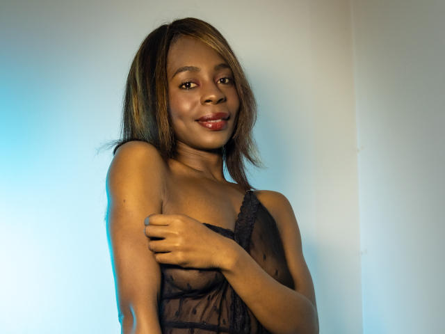 Angeiiina with dark brown skin, small breasts, and a black lace top, smiling, touching her left arm, light blue and white background, straight brown hair with blonde highlights.