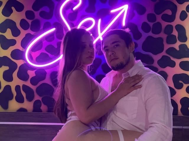 TinaYEdward with long brown hair, small tits, and white strappy lingerie, sits on a guy's lap, hand on his chest, wearing a white shirt, neon purple background.
