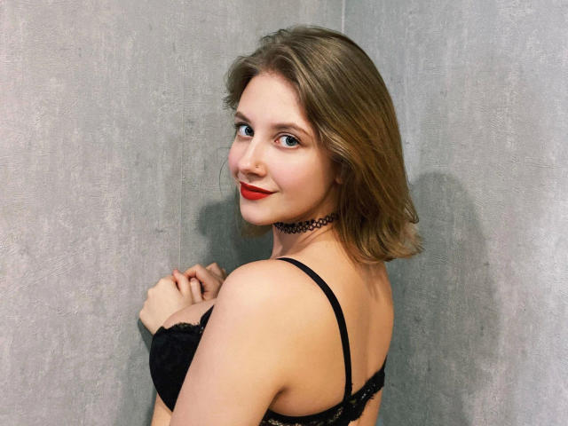 JuliaLuxx-hot with red lipstick, black bra, and choker, looking over her shoulder, smiling, her hands near her tits, brown hair, gray wall in the background.