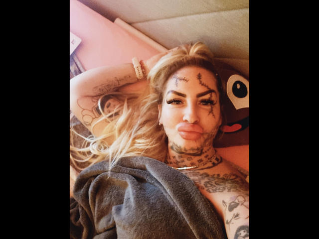 Jeanny-Bln-hot sdraiata su un letto, capelli biondi, tatuaggi sul viso e sul collo, eyeliner, piercing al naso, labbra piene, coperta da una coperta grigia, braccia dietro la testa, che guarda la fotocamera..