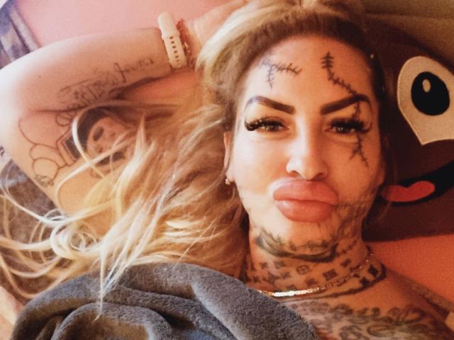 Jeanny-Bln, attraente con capelli biondi, tatuaggi sul viso, piercing al naso e labbra piene, sdraiata, con tatuaggi sul collo e sul braccio, una coperta grigia e una parete rosa sullo sfondo..