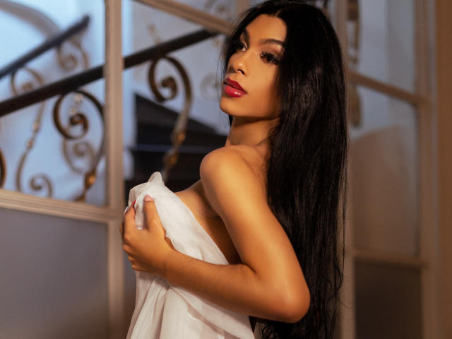 CelesteSweety mit langen, schwarzen Haaren, die ein weißes Handtuch um ihre Brüste trägt, hat roten Lippenstift und einen sexy, selbstbewussten Blick. Im Hintergrund sind Treppen mit einer verzierten Metallreling zu sehen..