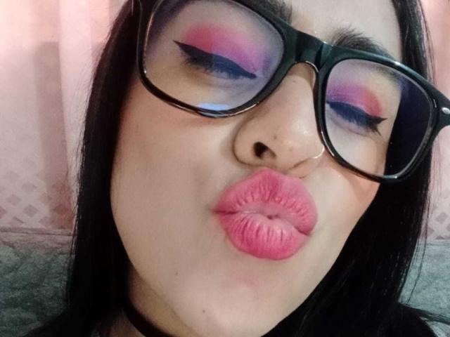 StellaBeckker's face with pink lipstick, puffy lips, black-framed glasses, and purple-blue eyeshadow, looking down with nose slightly pointed, wearing a black choker.