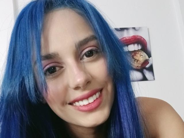 AnaisHilton with blue hair, smiling, showing teeth, pink eyeshadow, nose ring, and pink lipstick. Tongue ring visible. AnisHilton's tits not shown. Background has a lip photo.