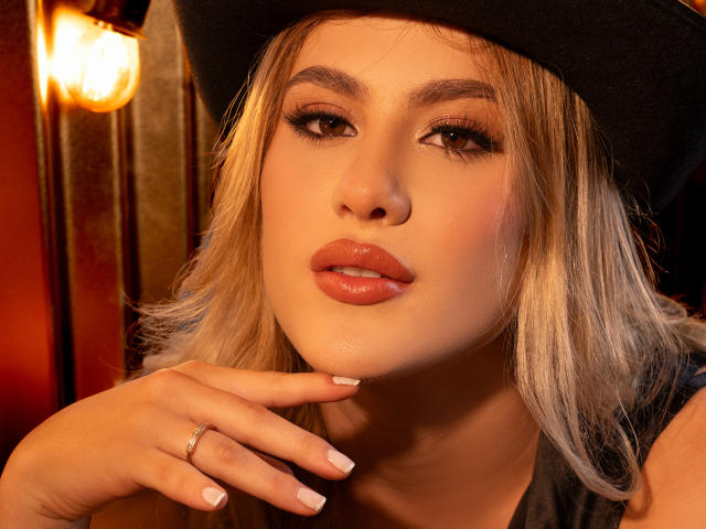 Adela Laurent, con le labbra piene, indossa un cappello nero, ha i capelli biondi e il trucco, e tocca il mento con le unghie curate, con un anello al dito, in una luce calda..