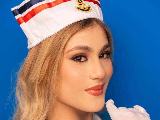 AdelaLaurent, blonde woman with wavy hair, wearing a sailor hat with red and blue stripes, white gloves, and orange lipstick, smiling, blue background, cute and sexy.