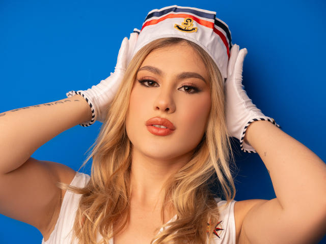 AdelaLaurent with blonde hair, white sailor hat, and gloves, pouting with orange lips, hands in her hair, wearing a white top, against a blue background.