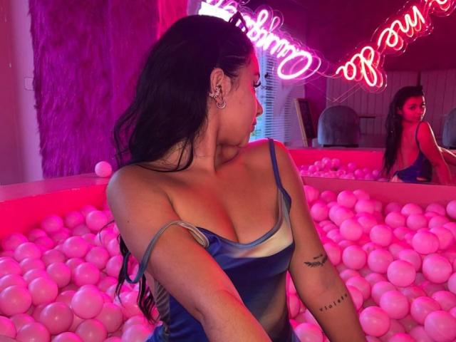 AlessiaFerry with long dark hair, wearing a blue and gray dress, showing her tits in a pink-lit bubble bath, tattoo on her right arm, looking to the side.