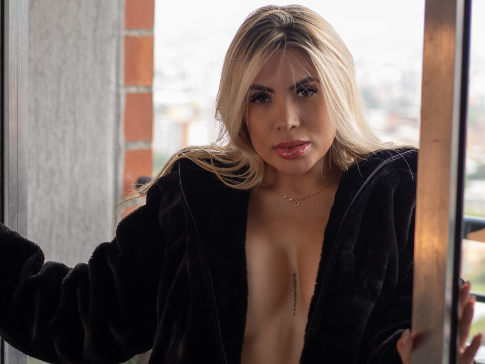 SaraMillees with blonde hair, wearing a black velvet robe, exposing her tits, a small vertical line tattoo between them, and a heart necklace, standing by a window.
