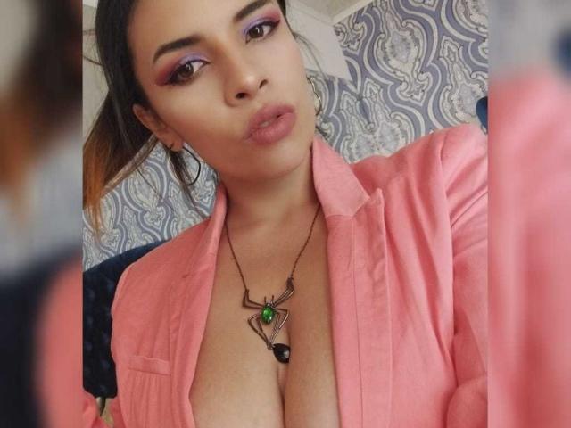 VioletaStone con una camisa rosa, abierta para mostrar un busto grande, sombra de ojos morada, collar verde, labios fruncidos, cabello castaño en una coleta, reflejada en el espejo..
