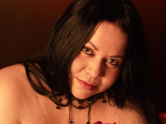 ValentinaFoxxy with black hair, green eyes, and a slight smile, wearing a black bead necklace, and a red accessory, has fair skin and is looking at the camera.