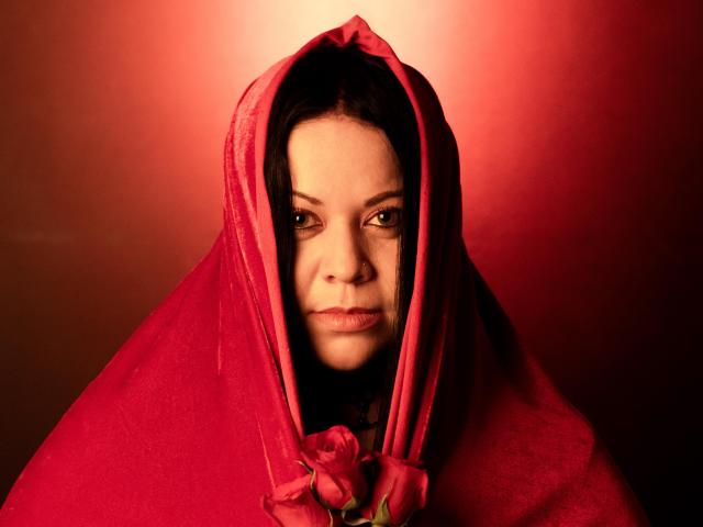 ValentinaFoxxy with red cloth covering her head and a rose in front, looking serious with a slight frown, red light in background. Her tits are partially visible.