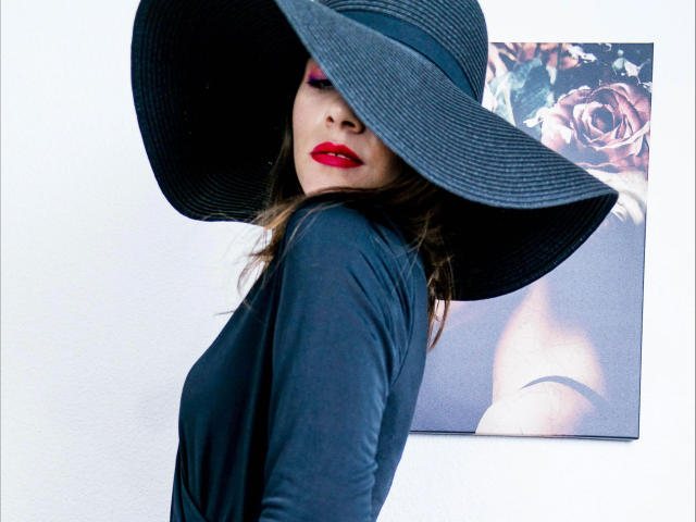 JustAlly-hot with red lipstick, black wide-brim hat, black long-sleeve shirt, looking over shoulder, brown hair, white wall, rose-themed poster, sexy, confident, stylish, attractive.