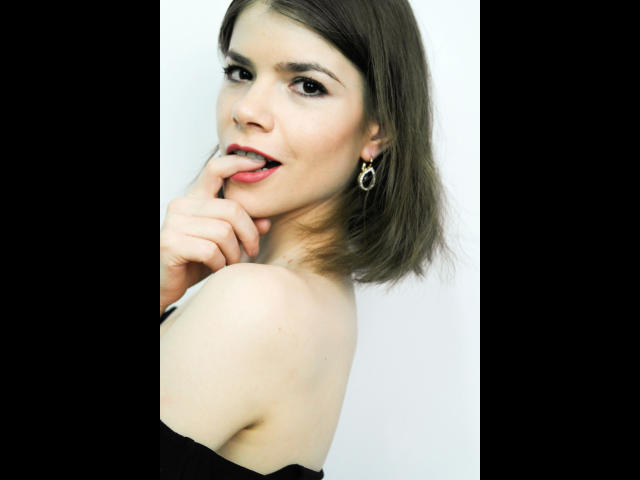 JustAlly-hot with short brown hair, biting her finger, looking at the camera, wearing a black strap, and gold earrings, showing her bare shoulder and slight smile.