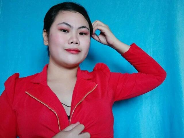 ChinitaPrincess con cabello oscuro, chaqueta roja con cremallera, mano en el cabello, fondo azul claro, sonrisa sutil, sombra de ojos rosa, lápiz labial rojo, busto medio, piel clara, pose segura..