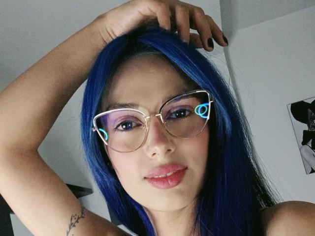 AnaisHilton with blue hair, wearing glasses with blue heart-shaped lenses, black nail polish, and a tattoo on her arm, smiling at the camera.