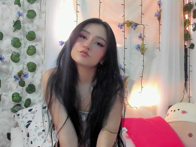 Melanniexcute with long black hair, pink eyeliner, and a cross necklace, pouting with slightly parted lips, wearing a strapless top, sitting on a pink pillow.