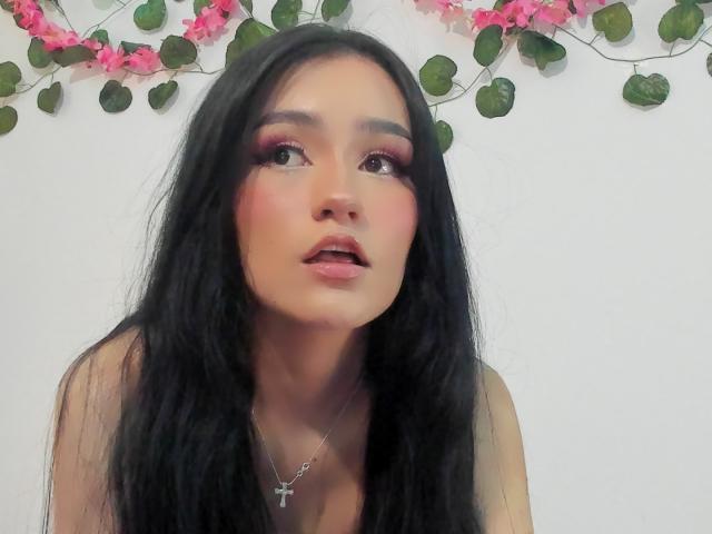 Melanniexcute with long black hair, pink eyeshadow, and pink lipstick, looking up with parted lips, wearing a silver cross necklace, her tits slightly visible, against a white background.