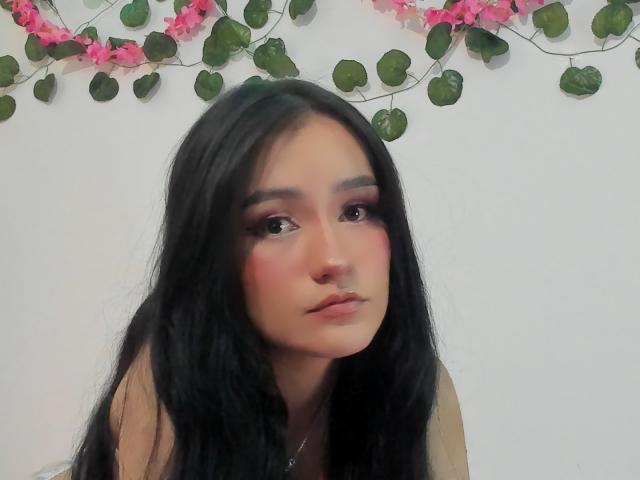Melanniexcute with long black hair, pinkish blush on cheeks, brown eyes, and slight pout, wearing a strapless top, pink floral garland in background.