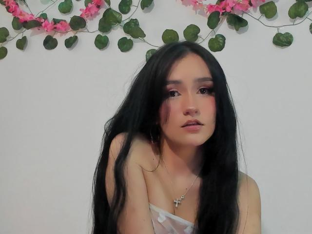 Melanniexcute with long black hair, pink eyeshadow, and a cross necklace, looking up with a slight pout, wearing a sheer white top, showing her small tits.