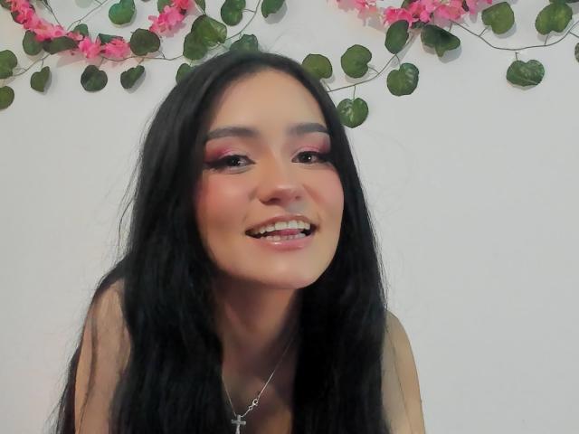 Melanniexcute with long black hair, pink eye makeup, and a smile, showing her shiny teeth, wearing a necklace, and looking cute. Her ass is not visible.