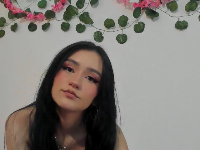Melanniexcute with long black hair, pink eye makeup, and pouty lips, looking at the camera. She has a cute face and is wearing a necklace, with pink floral vines.