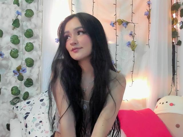 Melanniexcute with long black hair, pale skin, and subtle makeup, wearing a sleeveless top, smiling slightly, sitting on a bed with colorful pillows and fairy lights in the background.