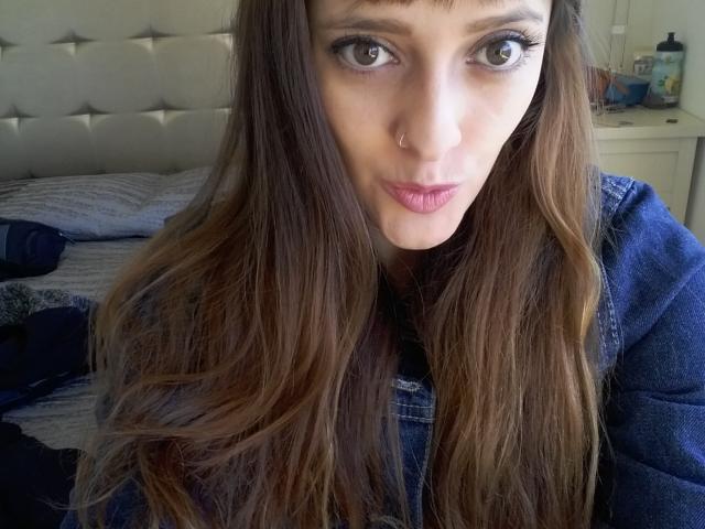 JaneRoa, avec de longs cheveux bruns, des yeux bleus et du rouge à lèvres rose, portant une veste en jean, regardant l'appareil photo avec une légère moue, dans une chambre bien éclairée..