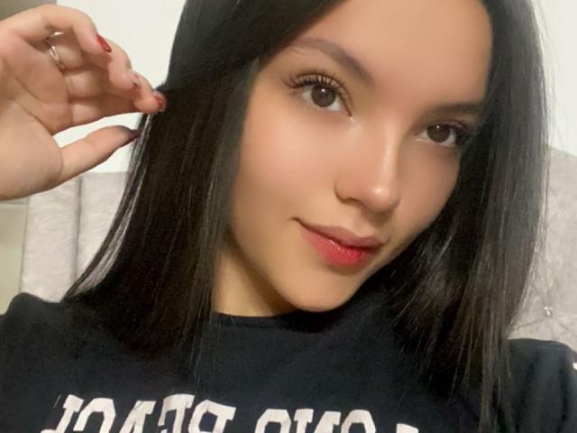 IssaPuasini has smooth brown skin, long black hair, and red lipstick, smiling with brown eyes, wearing a black shirt, touching her hair with red nails.