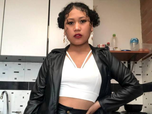 SophieSonyas with curly black hair, wearing a black leather jacket, white crop top, and black pants, standing in a kitchen, hands on hips, looking at the camera.