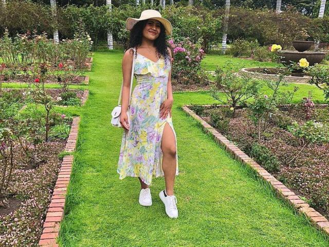 SophieSonyas with dark curly hair, wearing a sunhat, floral dress with a thigh slit, white sneakers, holding a white purse, smiling in a lush garden.